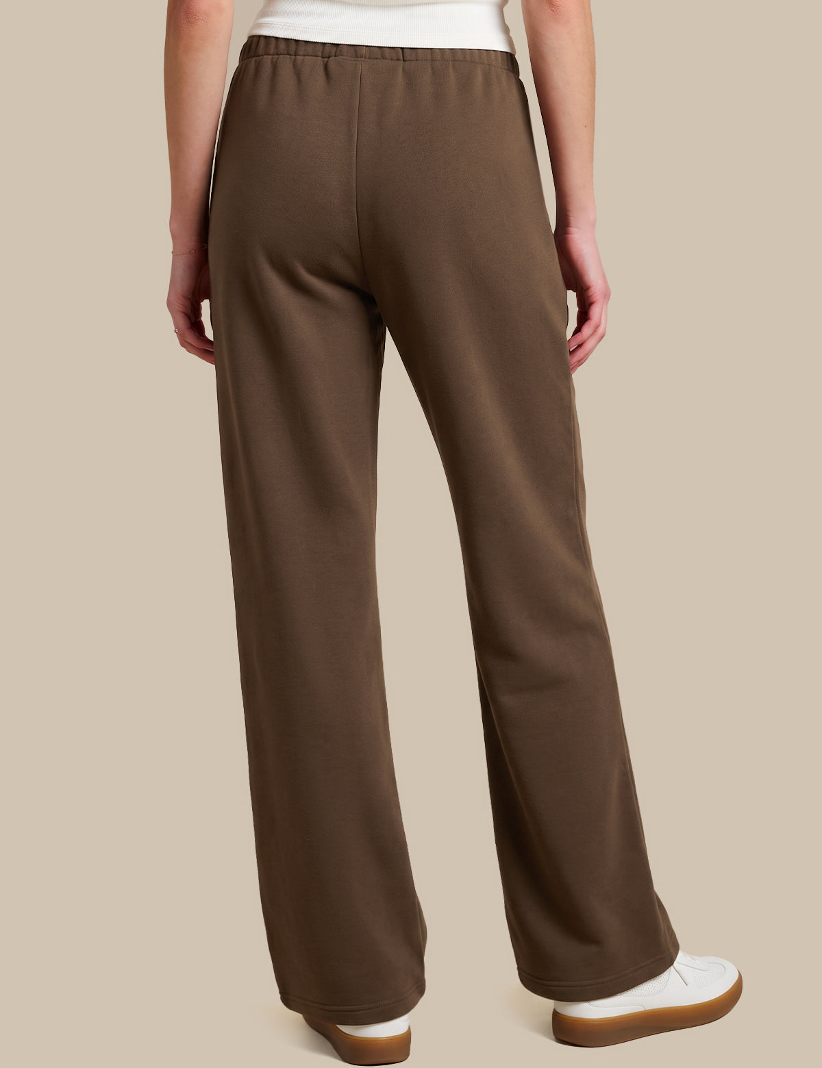 A person shown from the back wearing brown wide-leg pants, a white top, and white slip-on shoes against a plain beige studio background.