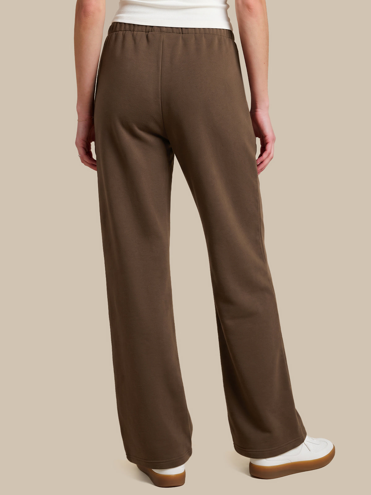 A person shown from the back wearing brown wide-leg pants, a white top, and white slip-on shoes against a plain beige studio background.