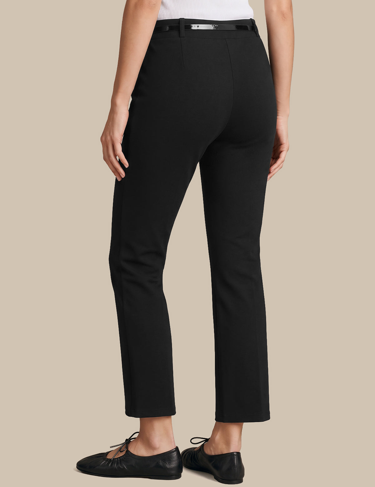 A model shown from the back wearing black cropped trousers with a matching black belt, paired with black leather flats, against a solid beige background.