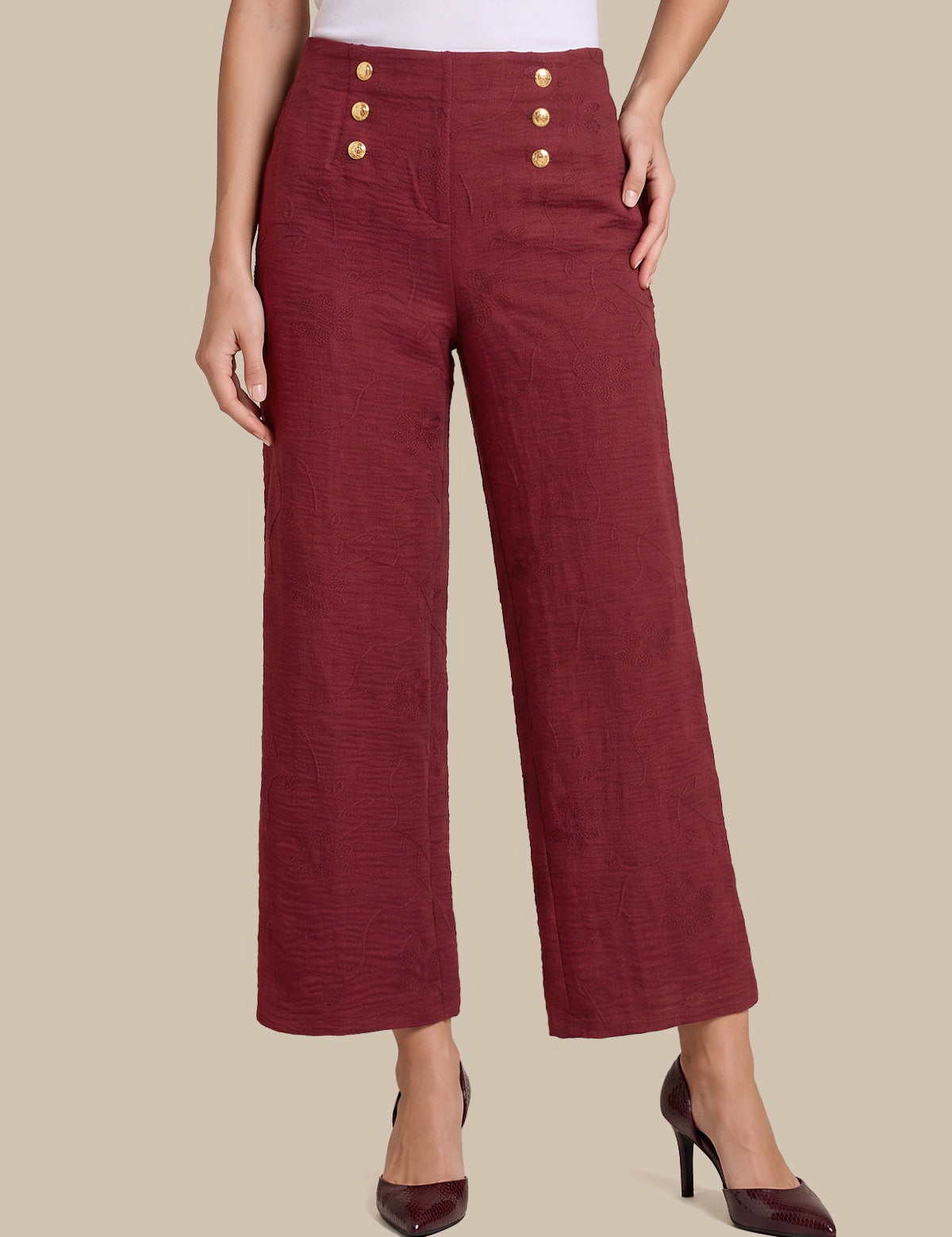 A model wearing high-waisted, wide-leg cropped trousers in a textured burgundy fabric, featuring decorative gold buttons on the front waistband, styled with dark burgundy high heels against a solid beige background.