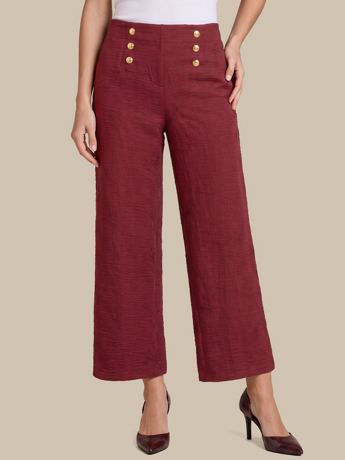 A model wearing high-waisted, wide-leg cropped trousers in a textured burgundy fabric, featuring decorative gold buttons on the front waistband, styled with dark burgundy high heels against a solid beige background.