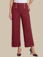 A model wearing high-waisted, wide-leg cropped trousers in a textured burgundy fabric, featuring decorative gold buttons on the front waistband, styled with dark burgundy high heels against a solid beige background.