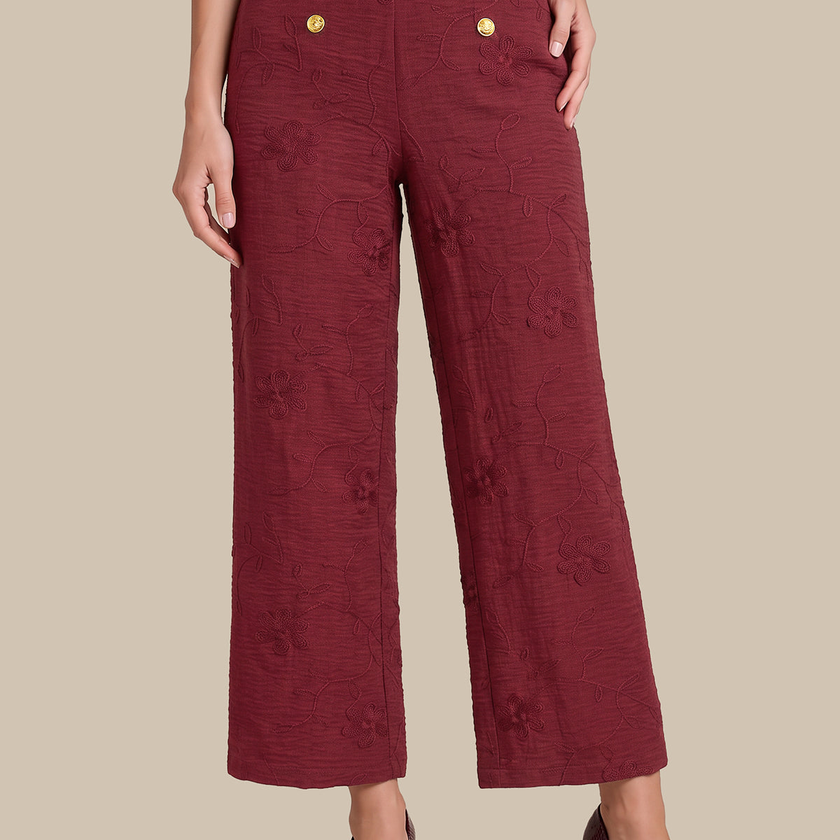 A model wearing high-waisted, wide-leg cropped trousers in a textured burgundy fabric with gold buttons on the front, paired with dark burgundy heels, against a solid beige background.