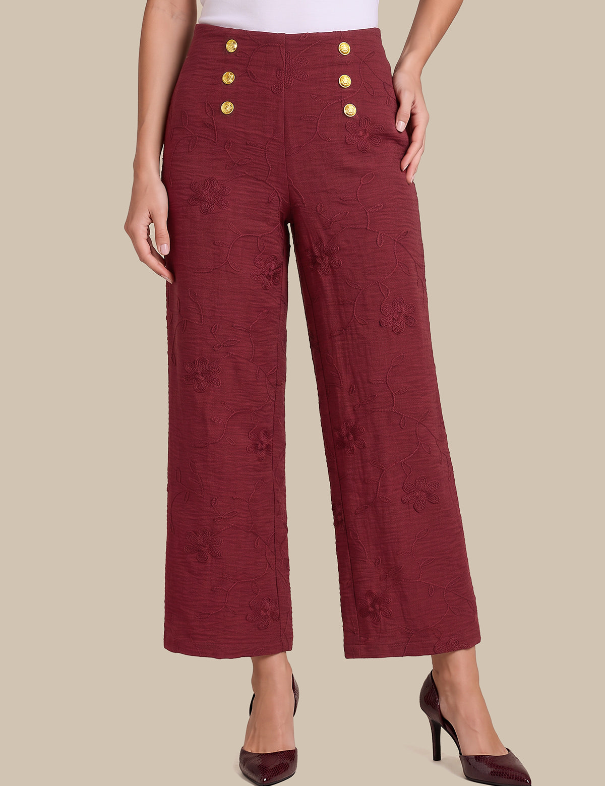 A model wearing high-waisted, wide-leg cropped trousers in a textured burgundy fabric with gold buttons on the front, paired with dark burgundy heels, against a solid beige background.