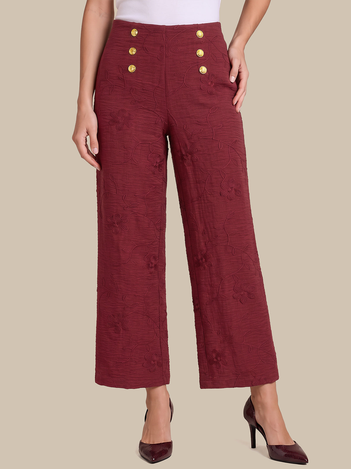 A model wearing high-waisted, wide-leg cropped trousers in a textured burgundy fabric with gold buttons on the front, paired with dark burgundy heels, against a solid beige background.