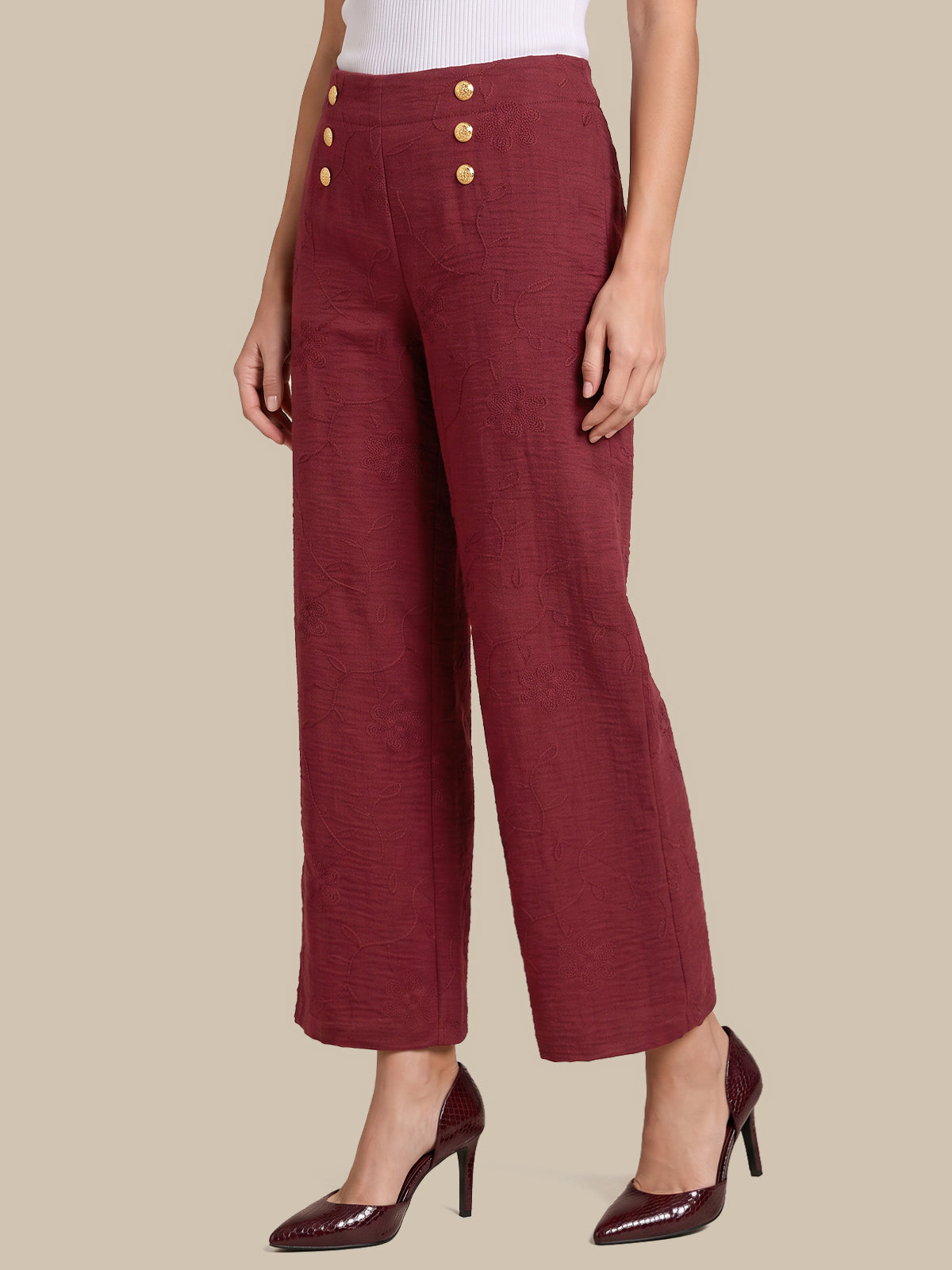 A model is shown from the waist down wearing high-waisted, wide-leg cropped trousers in a textured burgundy fabric with gold buttons on the front, paired with burgundy high heels against a solid beige background.