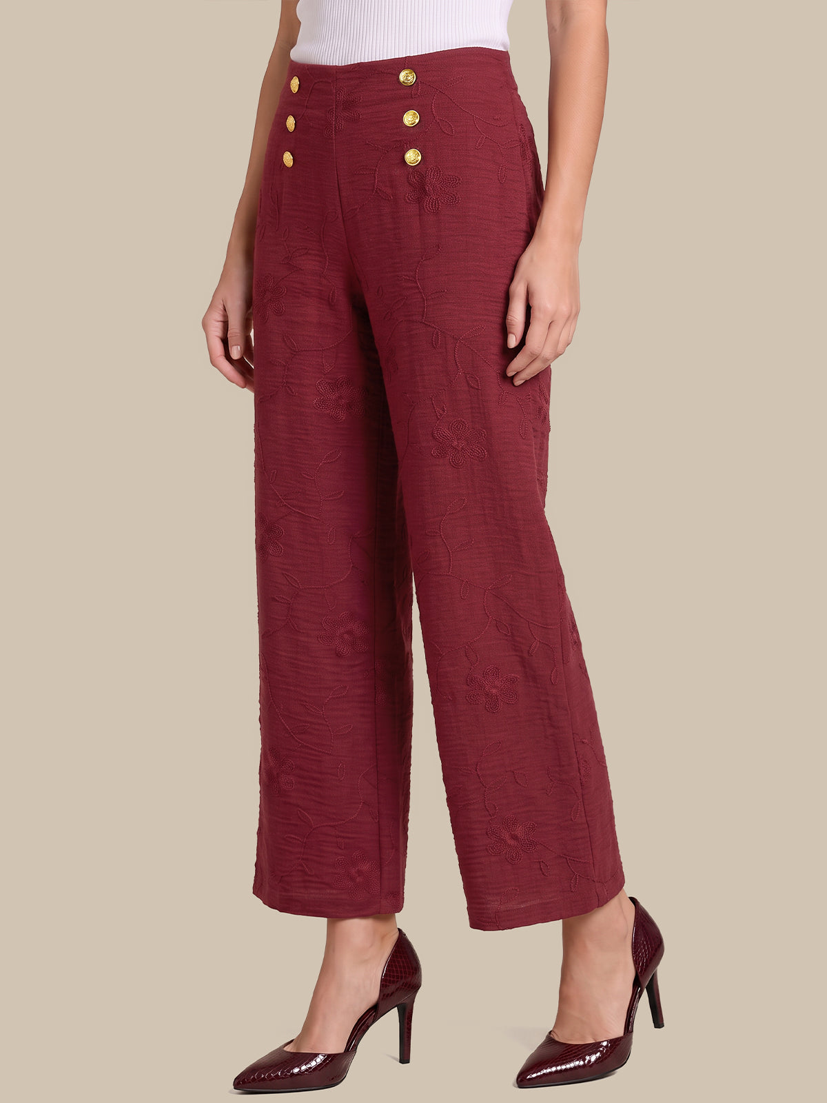 A model is shown from the waist down wearing high-waisted, wide-leg cropped trousers in a textured burgundy fabric with gold buttons on the front, paired with matching burgundy high heels against a solid beige background.