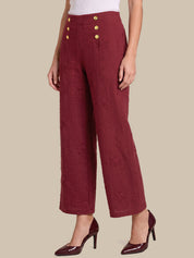 A model is shown from the waist down wearing high-waisted, wide-leg cropped trousers in a textured burgundy fabric with gold buttons on the front, paired with matching burgundy high heels against a solid beige background.