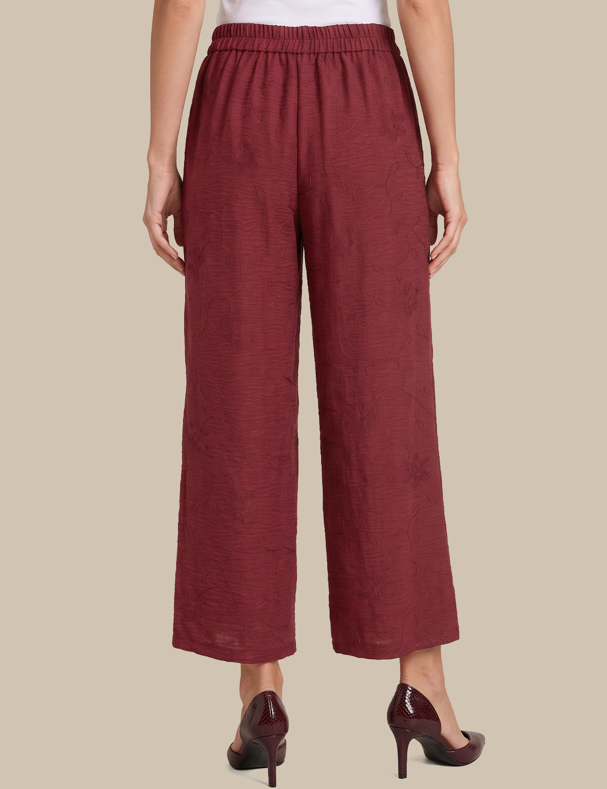 A model is shown from the back wearing a pair of wide-leg cropped pants in a textured burgundy fabric, paired with dark burgundy high heels against a solid beige background.