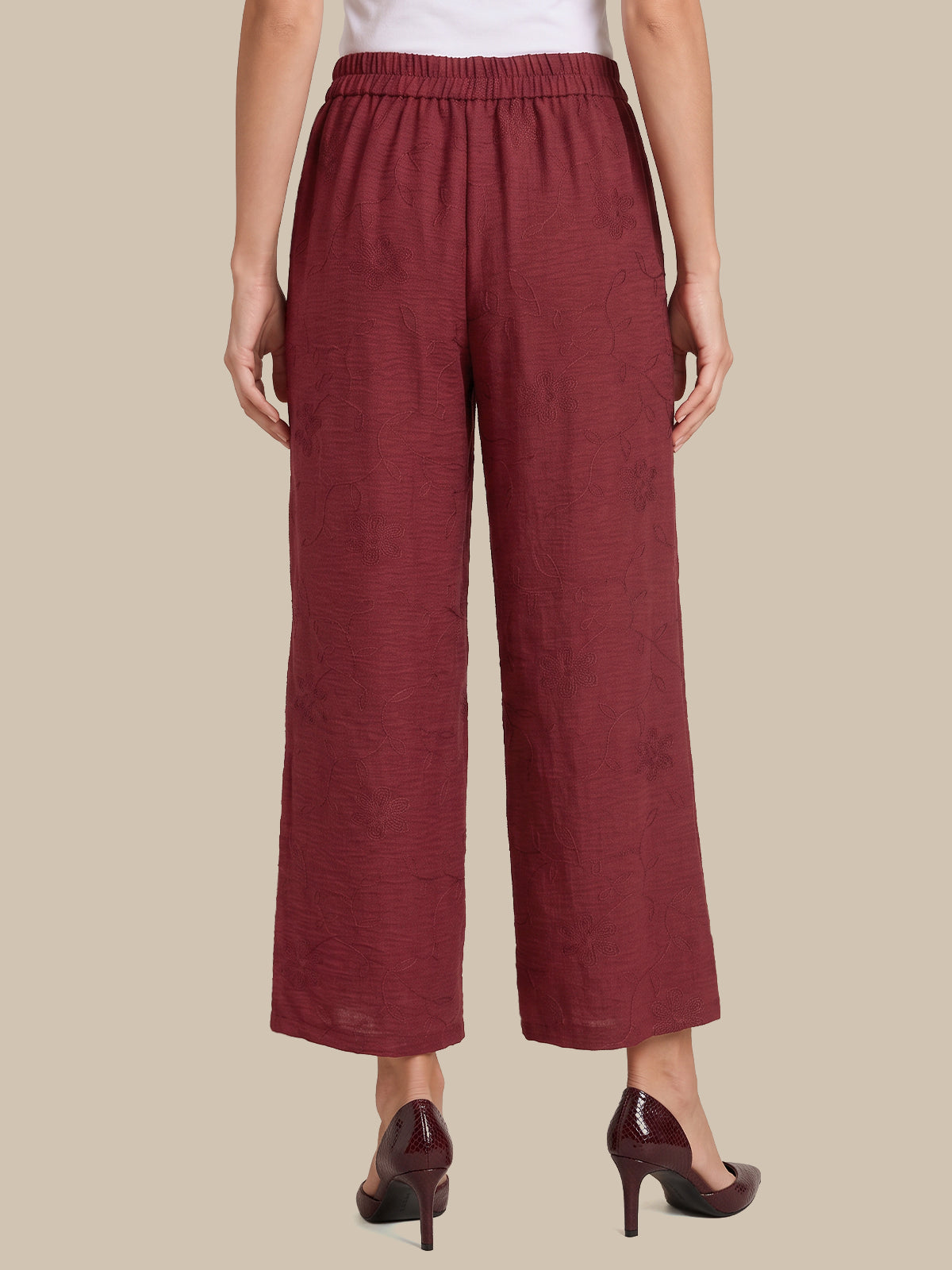 A model is shown from the back wearing a pair of wide-leg cropped pants in a textured burgundy fabric, paired with dark burgundy high heels against a solid beige background.