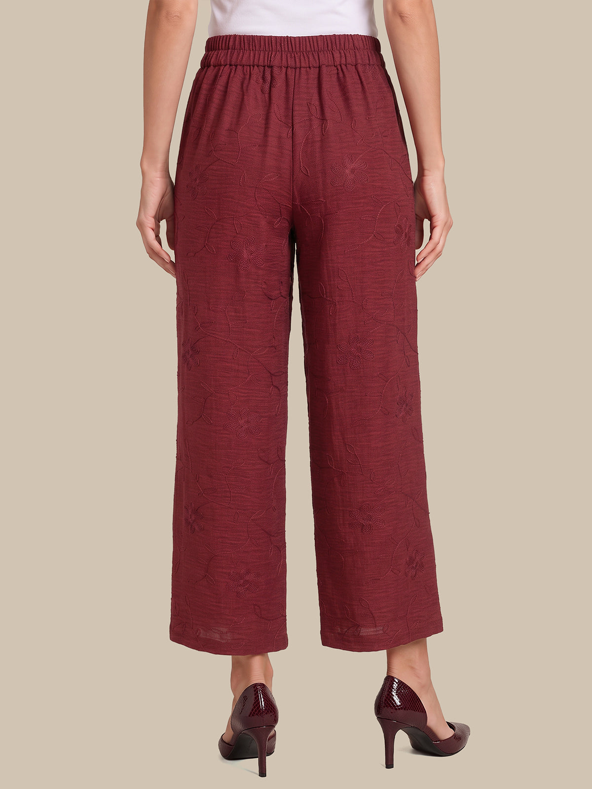 A person shown from the back wearing loose, wide-leg cropped pants in a deep burgundy tone with a subtle textured pattern, paired with dark pointed-toe heels against a plain beige studio background.