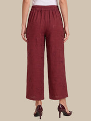 A person shown from the back wearing loose, wide-leg cropped pants in a deep burgundy tone with a subtle textured pattern, paired with dark pointed-toe heels against a plain beige studio background.