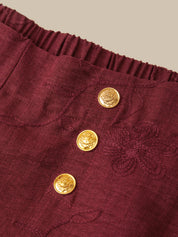 Close-up of a burgundy textured fabric garment featuring a gathered elastic waistband and three decorative gold buttons with floral engravings.