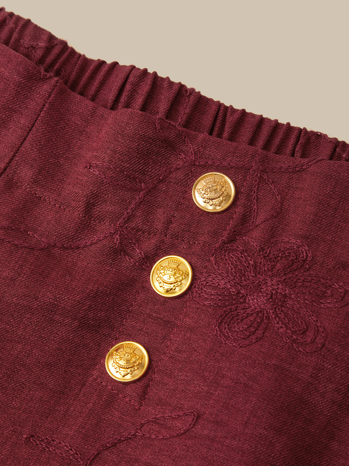 Close-up of a burgundy textured fabric garment featuring a gathered elastic waistband and three decorative gold buttons with floral engravings.