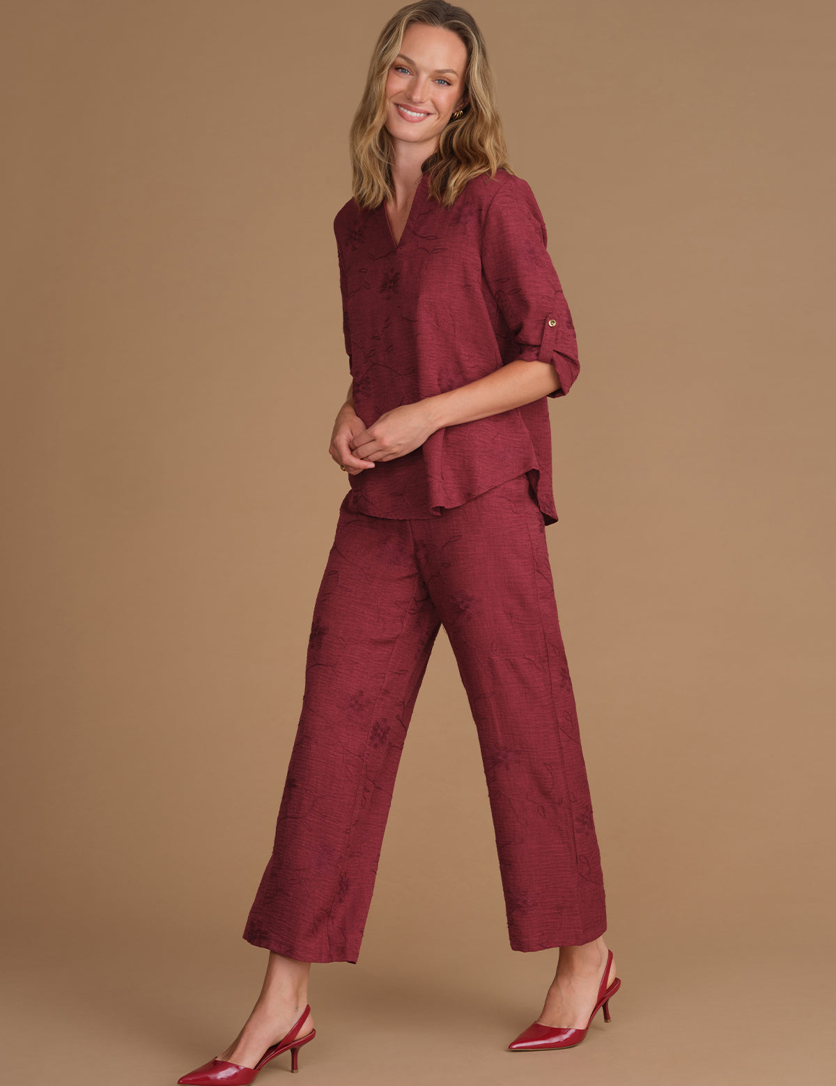 A woman modeling a matching burgundy two-piece set consisting of a textured short-sleeve button-up shirt and wide-leg cropped pants, paired with burgundy high heels, against a solid tan studio background.