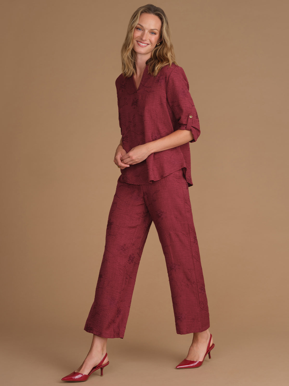 A woman modeling a matching burgundy two-piece set consisting of a textured short-sleeve button-up shirt and wide-leg cropped pants, paired with burgundy high heels, against a solid tan studio background.