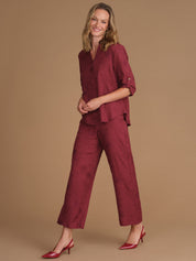A woman modeling a matching burgundy two-piece set consisting of a textured short-sleeve button-up shirt and wide-leg cropped pants, paired with burgundy high heels, against a solid tan studio background.