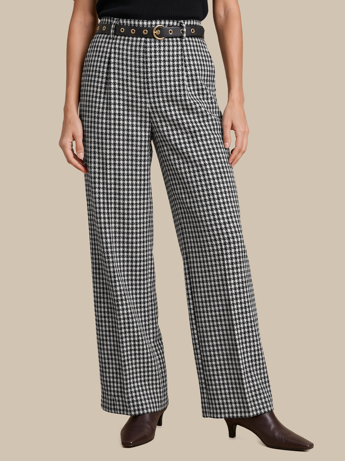 A model wearing high-waisted, wide-leg trousers in a black and white houndstooth pattern, styled with a black belt, a black top, and dark brown pointed-toe heeled boots against a solid beige background.