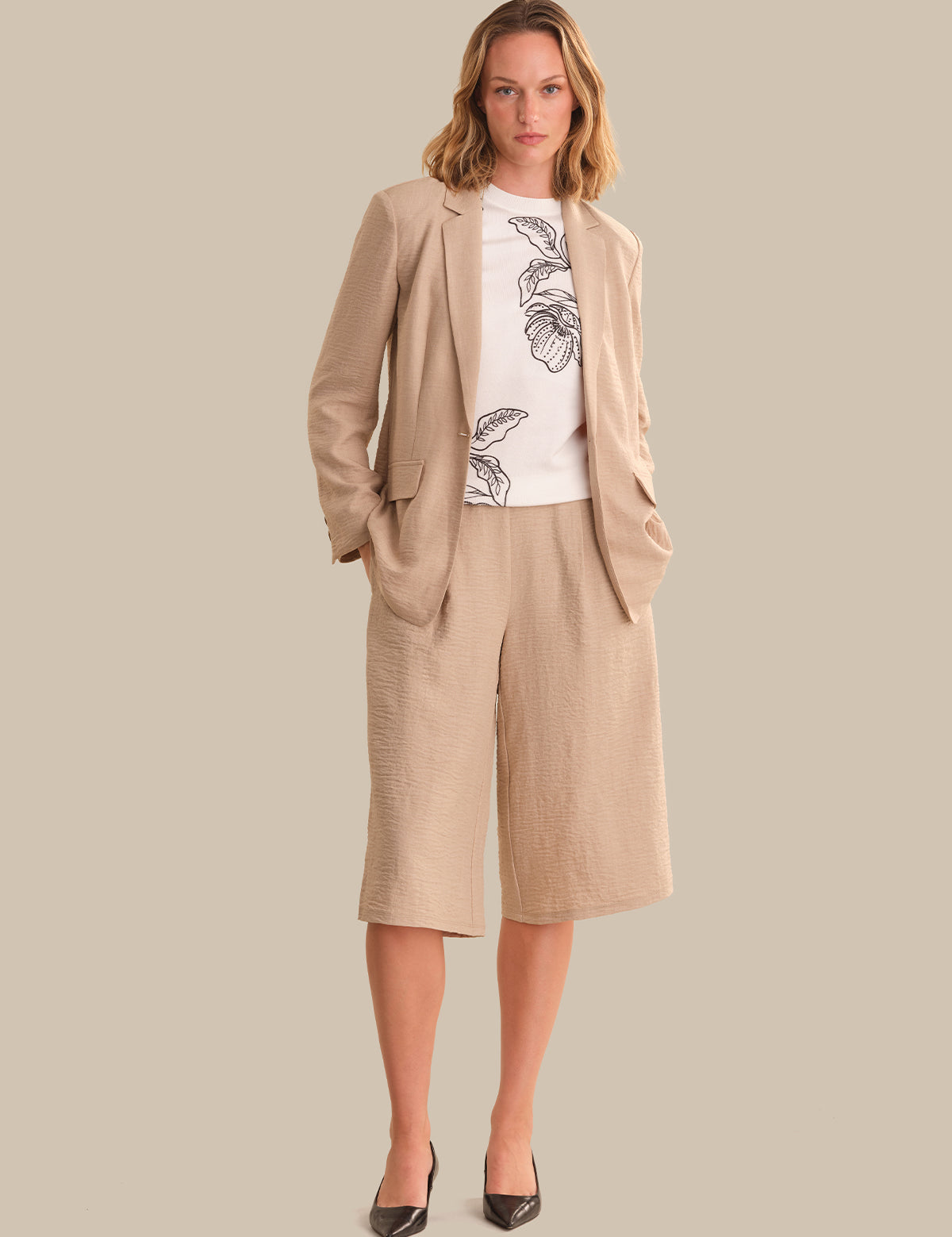 Woman wearing a beige suit with a white shirt featuring black floral patterns on a plain background