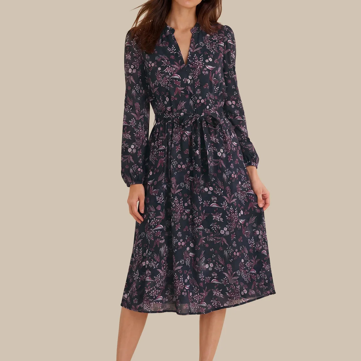 A woman modeling a long-sleeve midi dress with a dark navy and purple floral pattern, standing against a solid beige background.