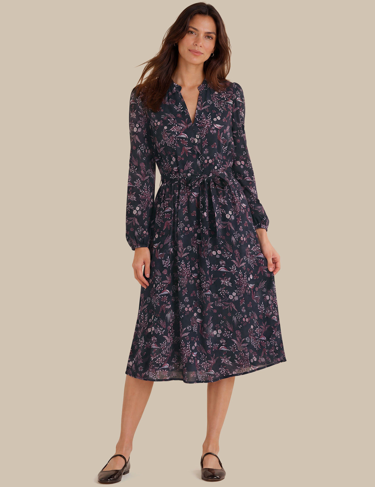 A woman modeling a long-sleeve midi dress with a dark navy and purple floral pattern, standing against a solid beige background.