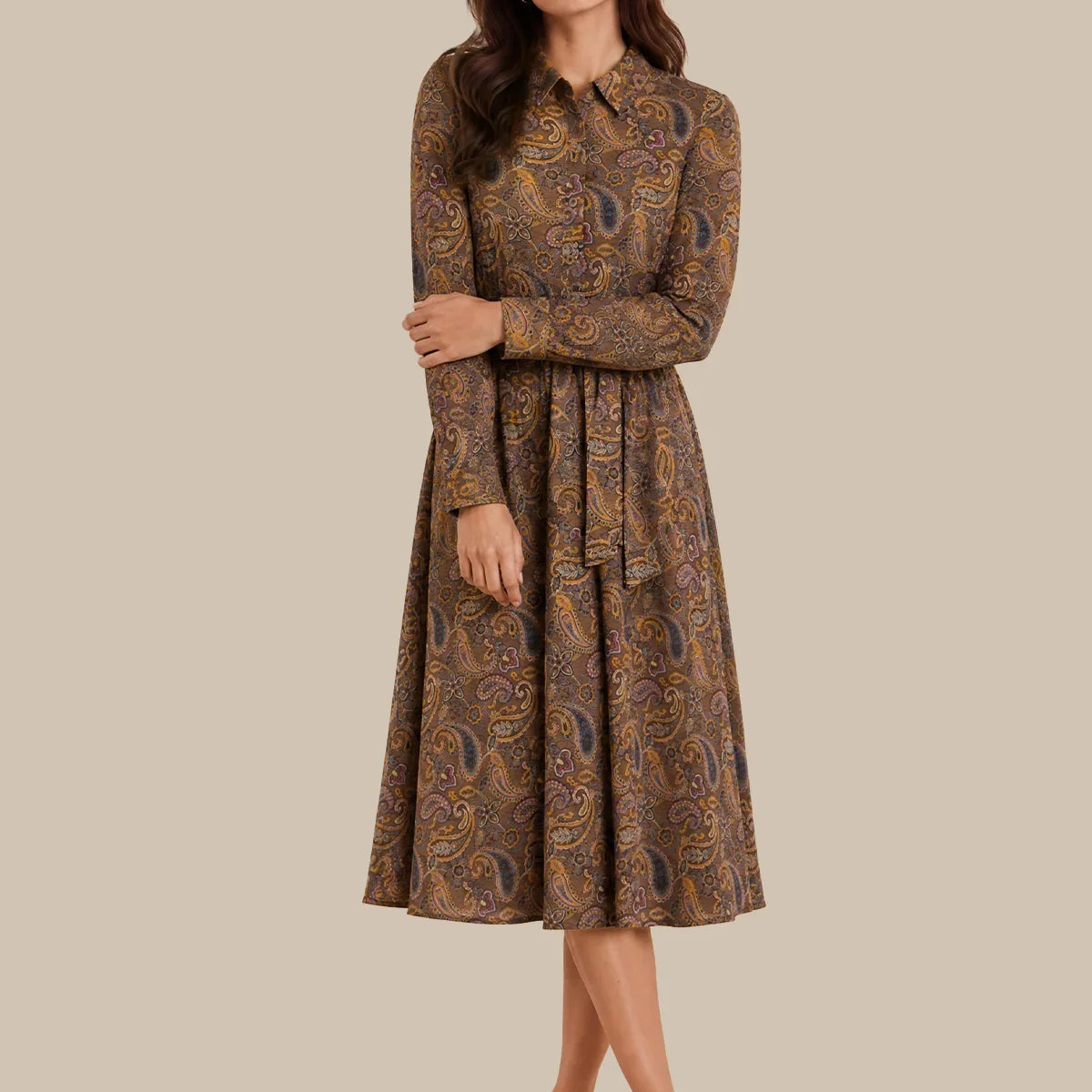 A woman modeling a long-sleeve midi dress with a brown and gold paisley pattern, standing against a solid beige background.