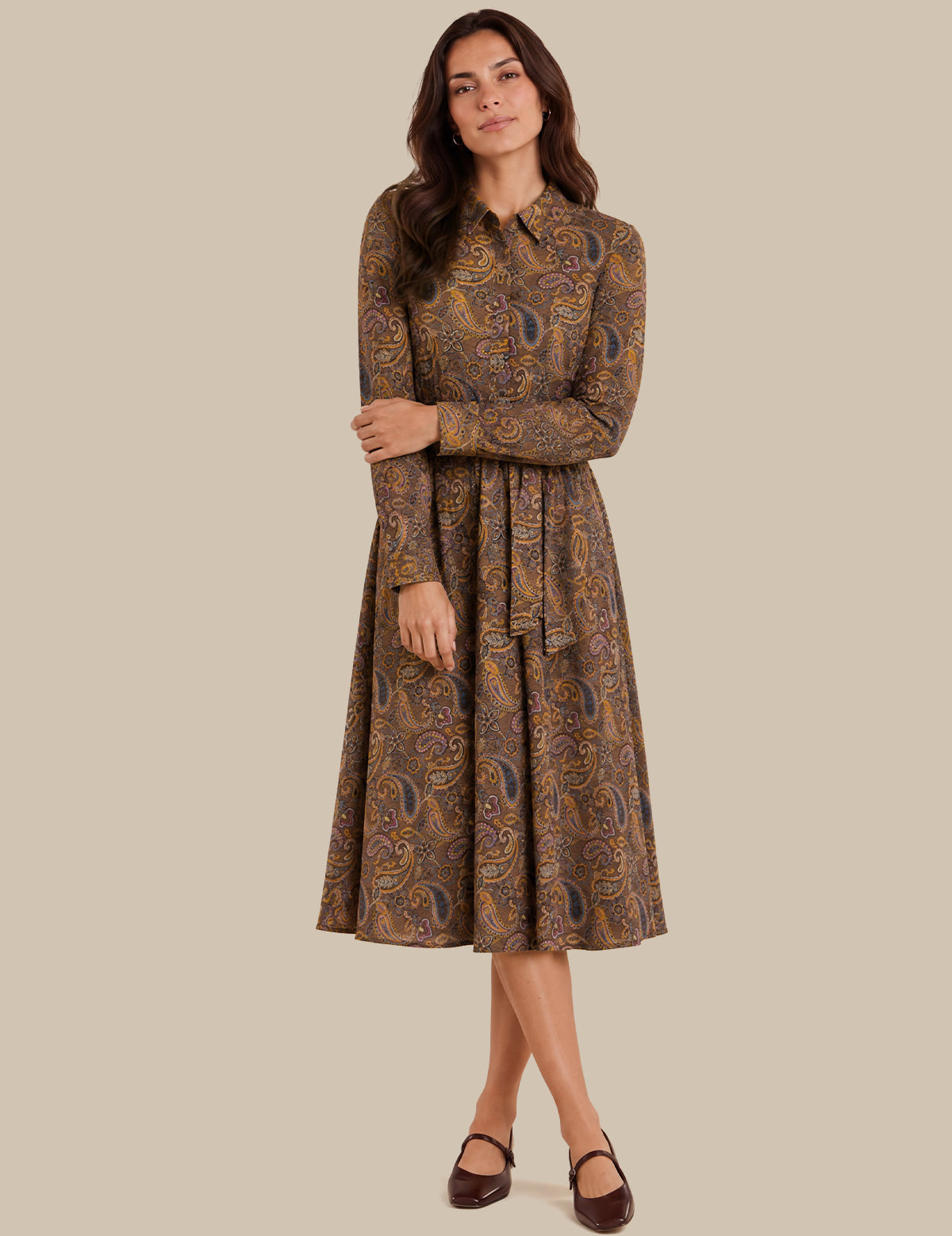 A woman modeling a long-sleeve midi dress with a brown and gold paisley pattern, standing against a solid beige background.