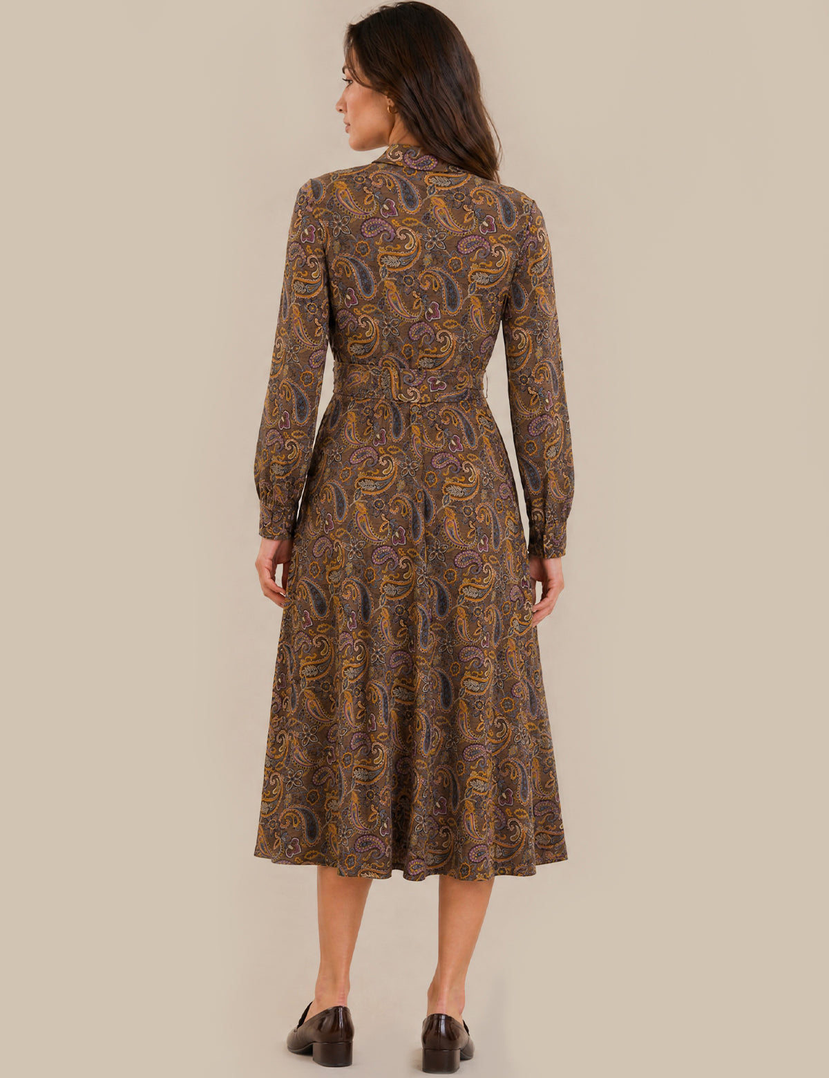 A woman viewed from the back wearing a long-sleeve midi dress with a brown, gold, and purple paisley pattern, paired with dark brown shoes, against a solid beige background.