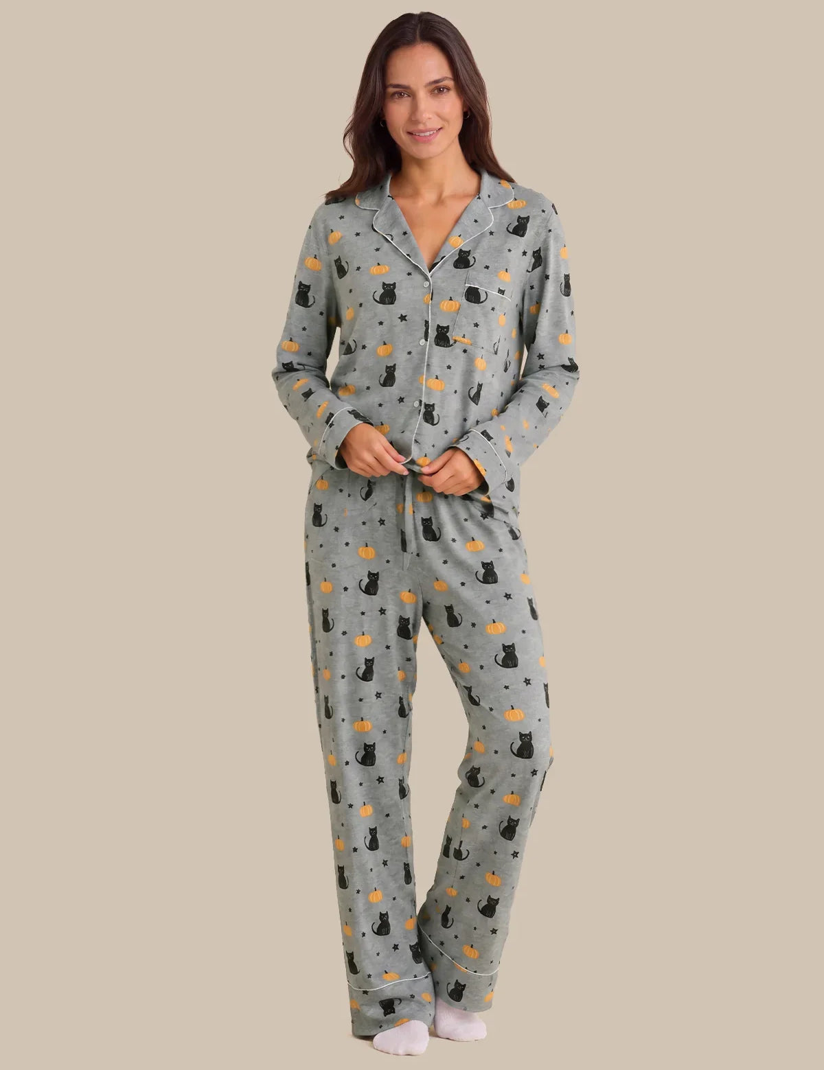 A woman modeling a two-piece pajama set with a gray base and an all-over print of black cats, white stars, and small orange celestial motifs, set against a solid beige background.