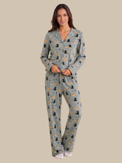 A woman modeling a two-piece pajama set with a gray base and an all-over print of black cats, white stars, and small orange celestial motifs, set against a solid beige background.