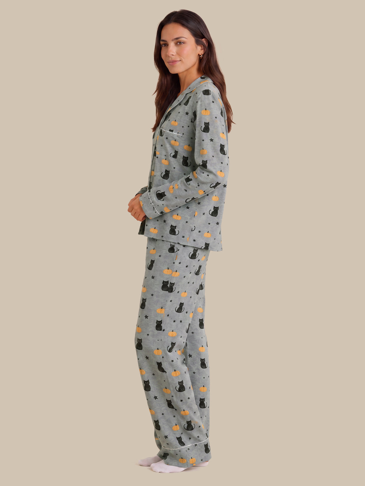 A woman modeling a two-piece pajama set with a light gray base and an all-over print of black cats, orange pumpkins, and small white dots, set against a solid beige background.