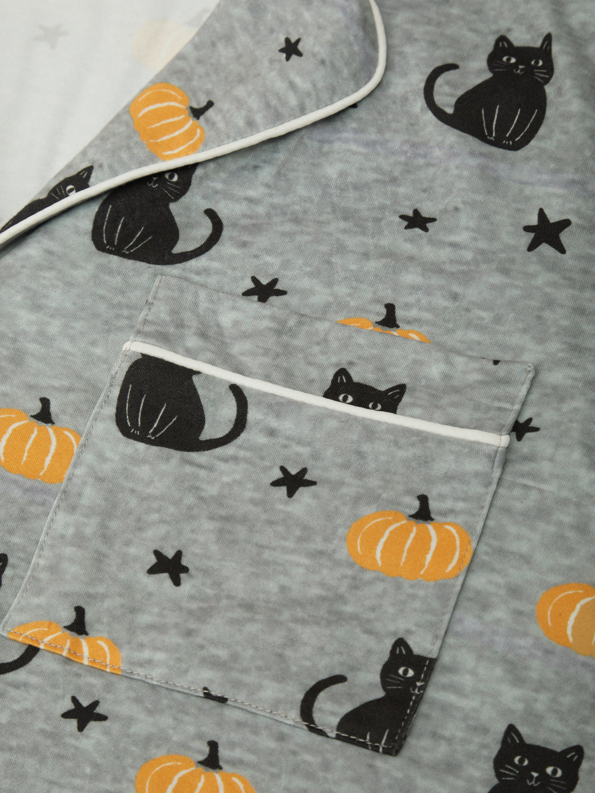 A close-up of a gray fabric item, likely a blanket or quilt, featuring a Halloween print of black cats, orange pumpkins, and black stars, with a white piping border around the edges and a front pocket.