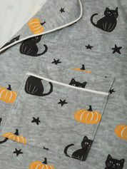 A close-up of a gray fabric item, likely a blanket or quilt, featuring a Halloween print of black cats, orange pumpkins, and black stars, with a white piping border around the edges and a front pocket.