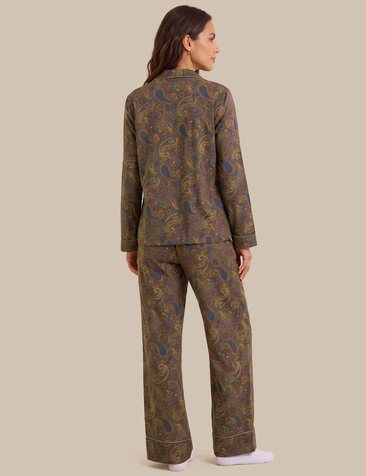 A woman shown from the back wearing a matching long-sleeve pajama set with an intricate paisley pattern in olive green, gold, and blue, paired with white socks, against a solid beige background.