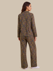 A woman shown from the back wearing a matching long-sleeve pajama set with an intricate paisley pattern in olive green, gold, and blue, paired with white socks, against a solid beige background.