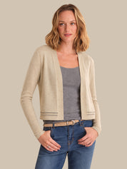 Woman wearing a beige cardigan over a gray top and blue jeans on a plain background