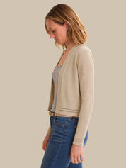 Woman wearing a beige cardigan over a gray shirt and blue jeans on a plain background