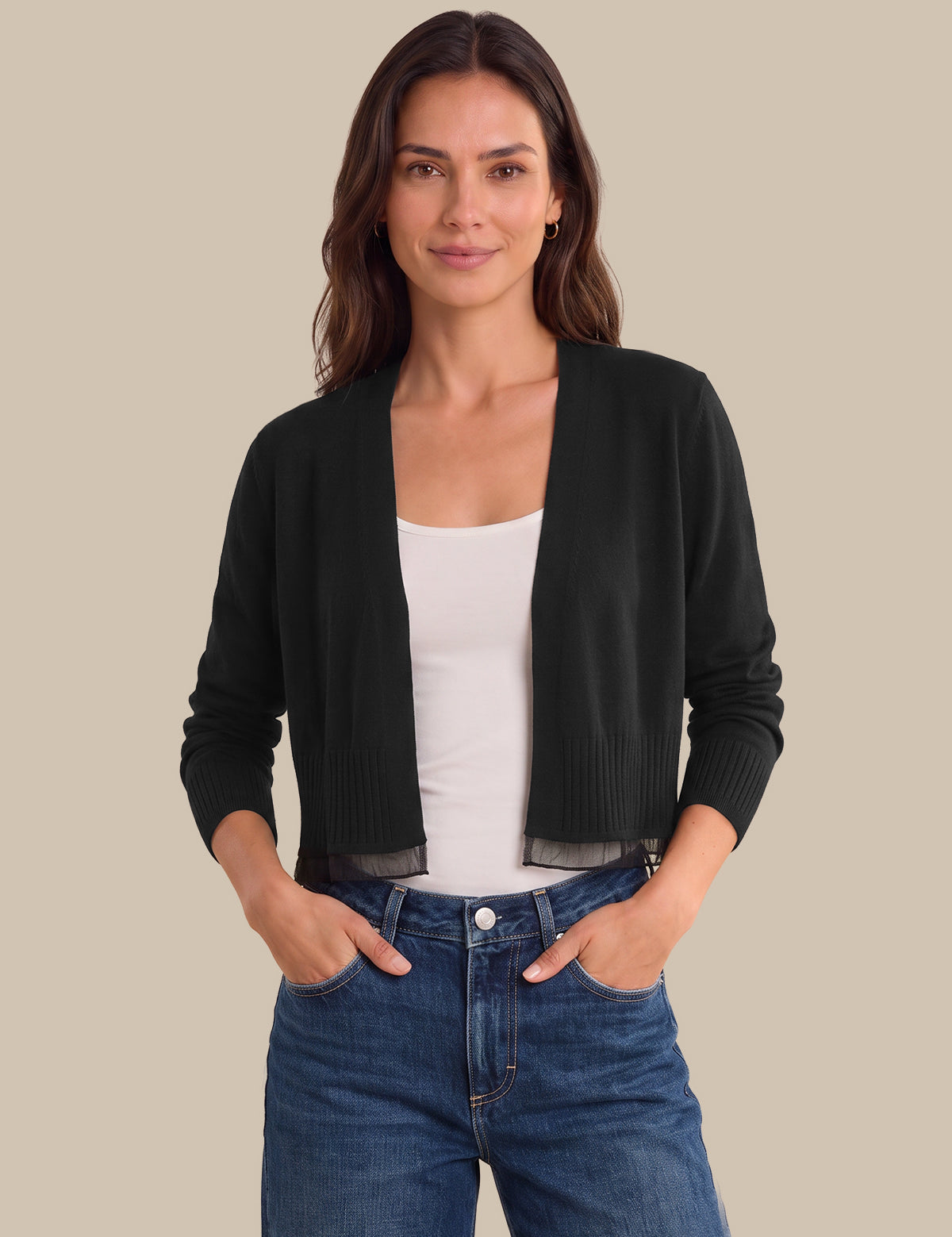 A woman modeling a black open-front knit cardigan over a white tank top and blue jeans, standing against a solid beige background.