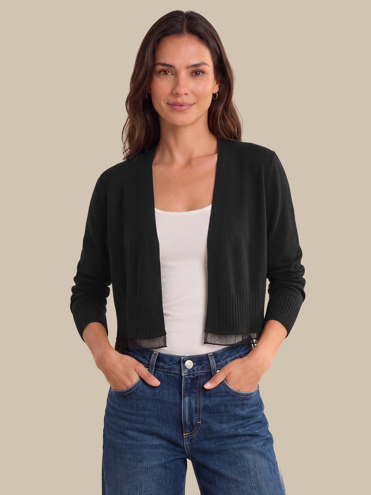 A woman modeling a black open-front knit cardigan over a white tank top and blue jeans, standing against a solid beige background.
