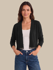 A woman modeling a black open-front knit cardigan over a white tank top and blue jeans, standing against a solid beige background.