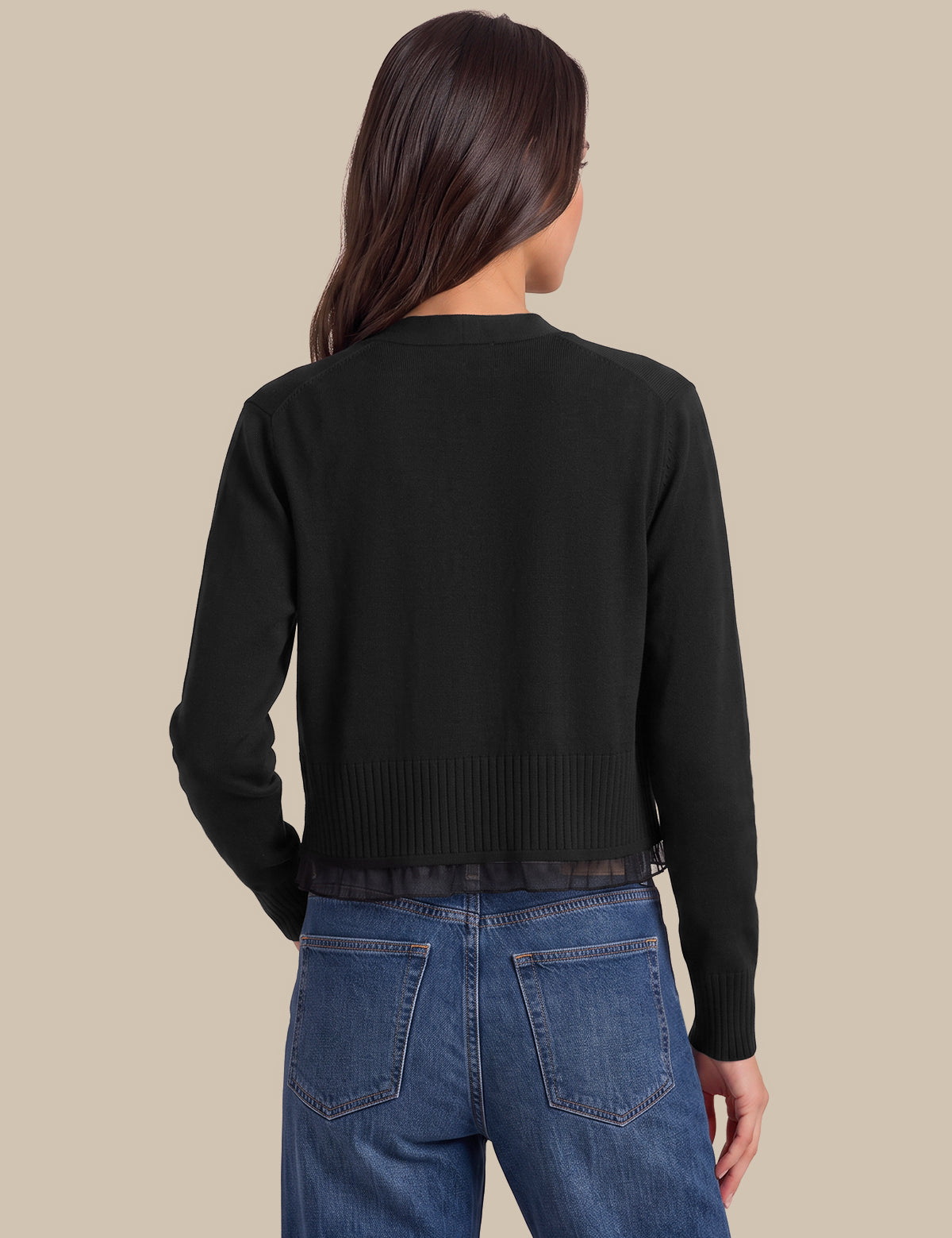 A woman viewed from the back wearing a black cropped knit cardigan with long sleeves and blue jeans, set against a solid beige background.