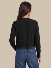 A woman viewed from the back wearing a black cropped knit cardigan with long sleeves and blue jeans, set against a solid beige background.