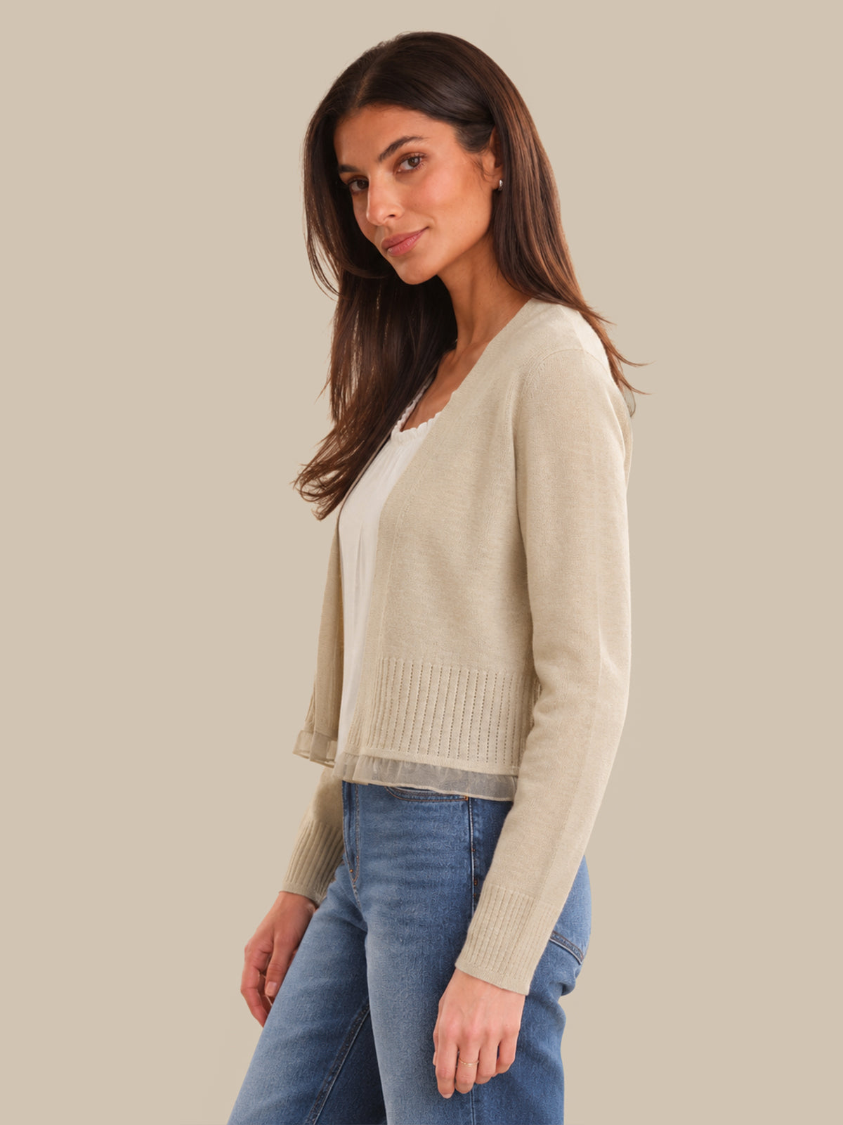 Woman wearing a beige cardigan over a white shirt with blue jeans on a beige background