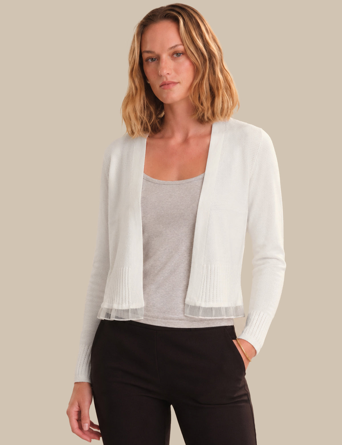 Woman wearing a white cardigan over a gray top and black pants on a beige background