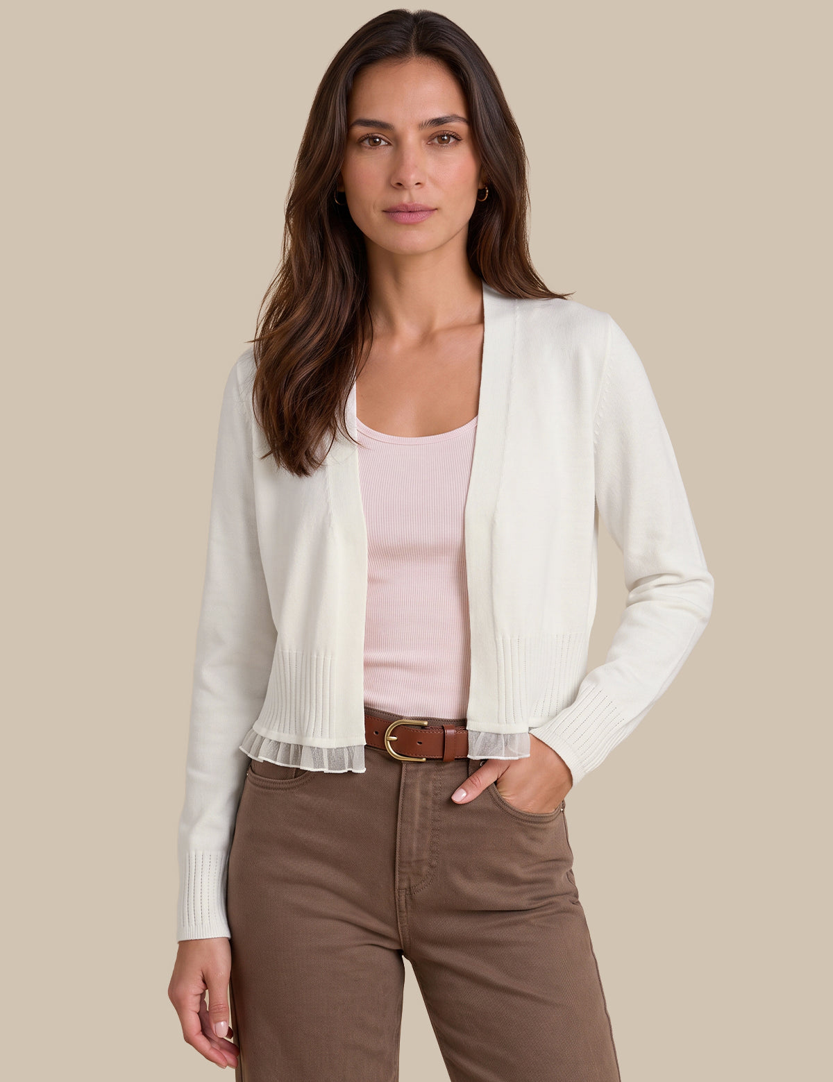A woman modeling a white ribbed knit cardigan over a light pink tank top, paired with brown pants and a brown belt, against a solid beige background.