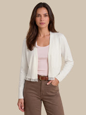 A woman modeling a white ribbed knit cardigan over a light pink tank top, paired with brown pants and a brown belt, against a solid beige background.
