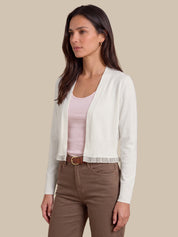 A woman modeling a white open-front knit cardigan with ruffle trim over a light pink top and brown pants, set against a solid beige background.