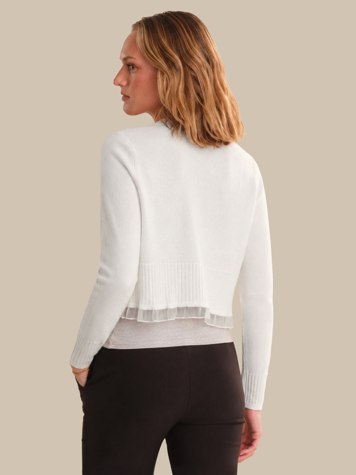 Woman wearing a white cardigan over a gray shirt with black pants on a beige background
