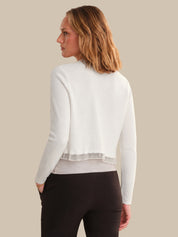 Woman wearing a white cardigan over a gray shirt with black pants on a beige background