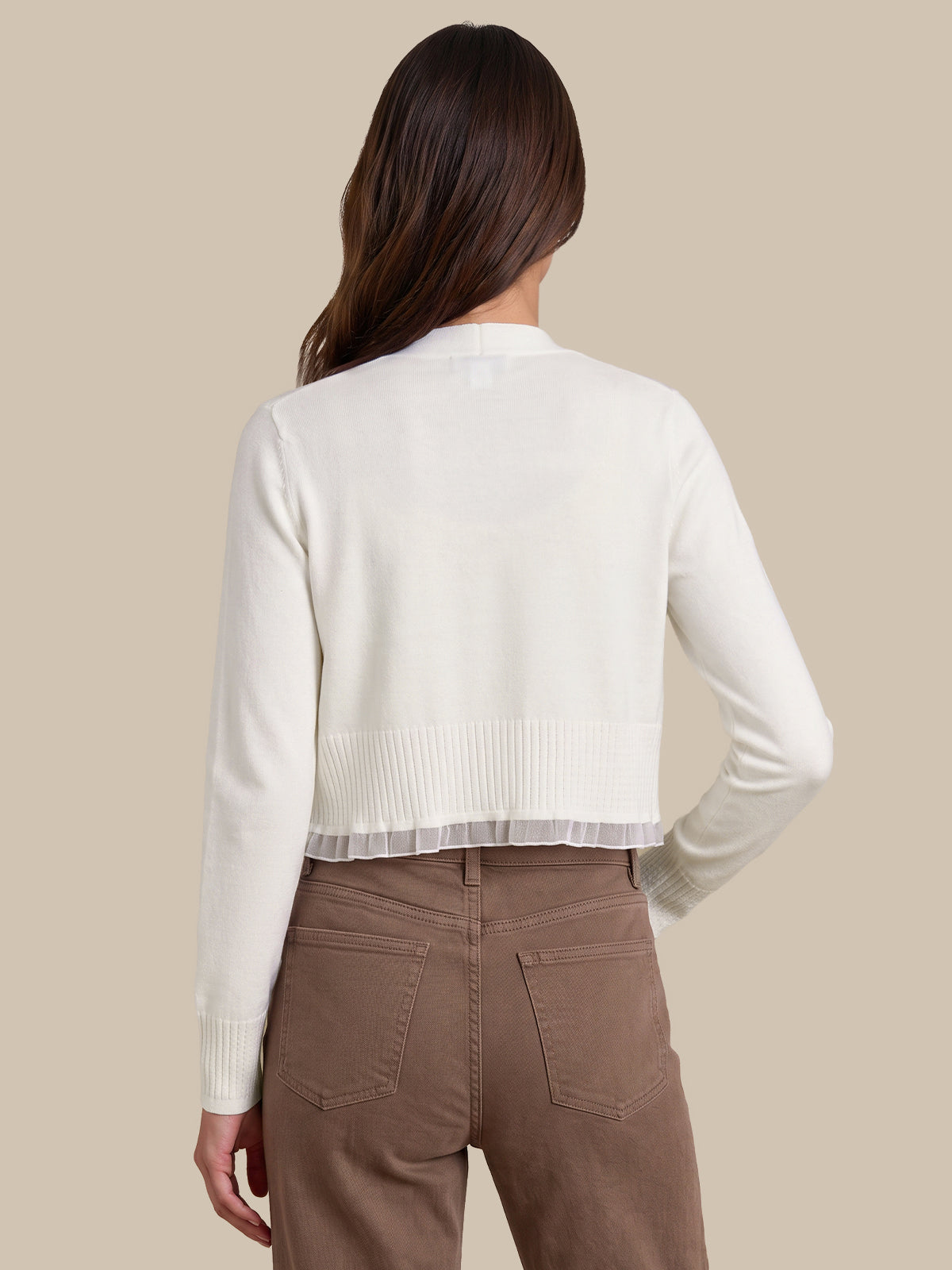 A woman shown from the back wearing a cream ribbed knit cropped cardigan with a sheer ruffle hem, paired with brown pants, against a solid beige background.
